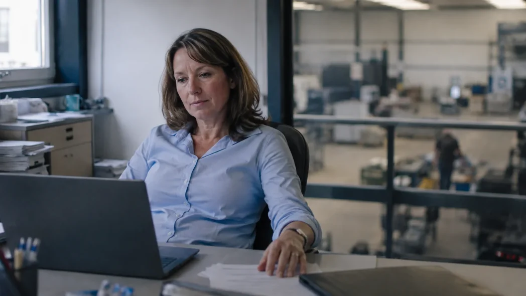Vrouw werkt achter een laptop op een kantoor met uitzicht op de werkplaats